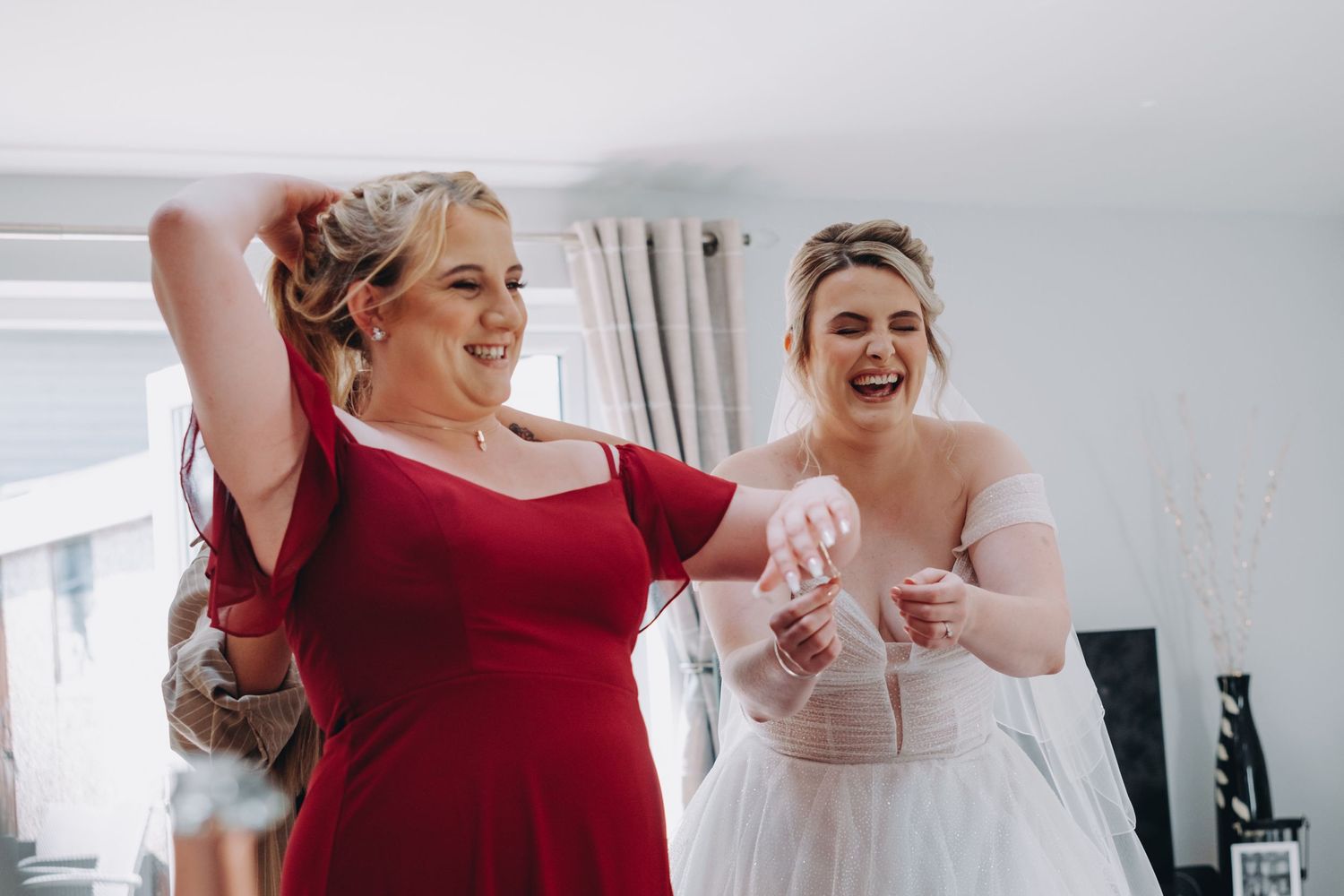 Bride and bridesmaid in dresses share a joyful laugh together while dancing at an indoor celebration.