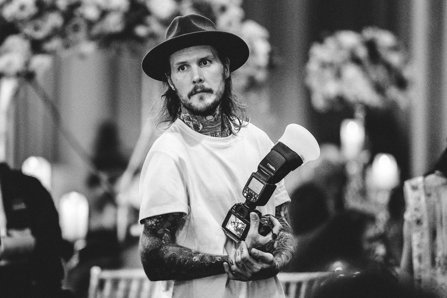 Black and white photo of photographer with tattoos holding camera wearing wide brim hat.