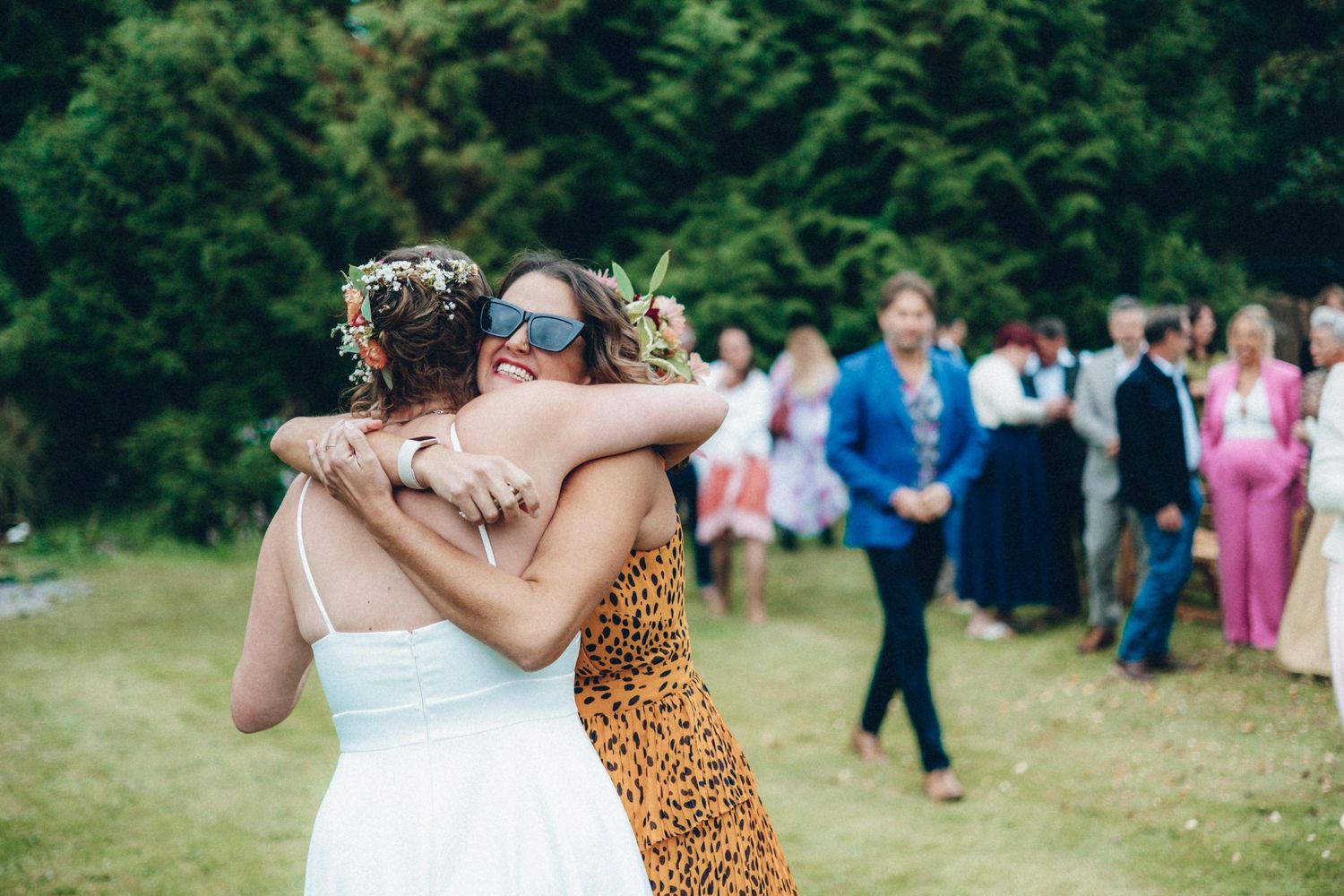 Two wedding guests embrace on a grassy lawn during the outdoor celebration.