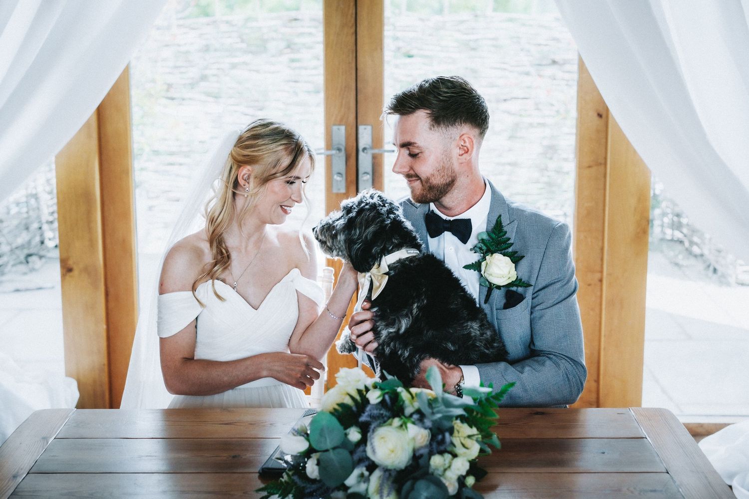A couple sits with their black dog at a wooden table under white drapes during their wedding celebration.