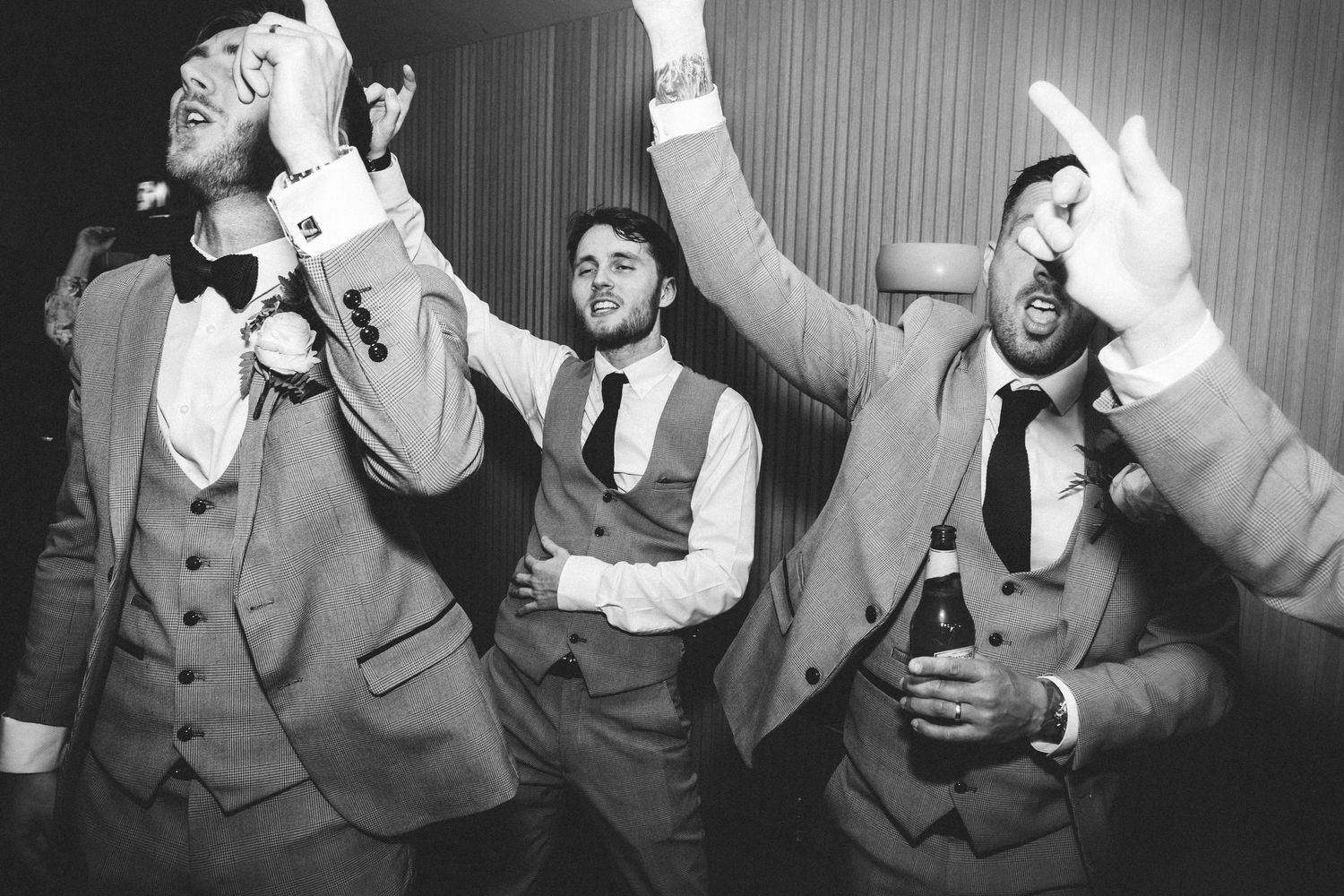 Groomsmen in formal attire dancing and celebrating at a wedding reception captured in black and white.