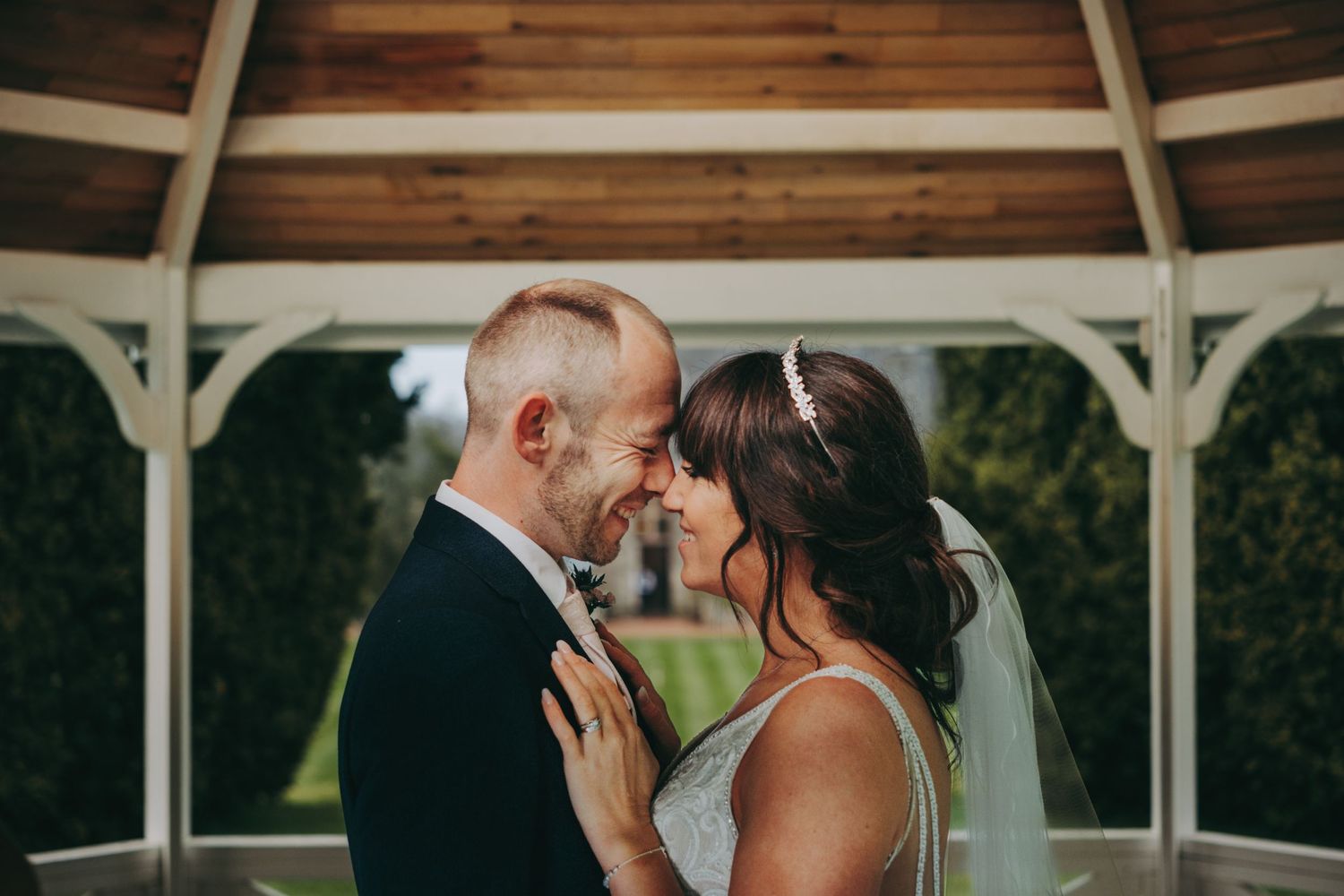 Couple shares romantic moment under large black umbrella on manicured lawn during rainy wedding day.