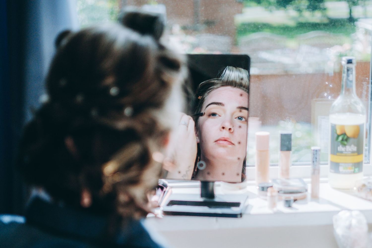 A reflection in a mirror shows a bridesmaid sitting at a table with a bottle and glassware in soft, blurry lighting.