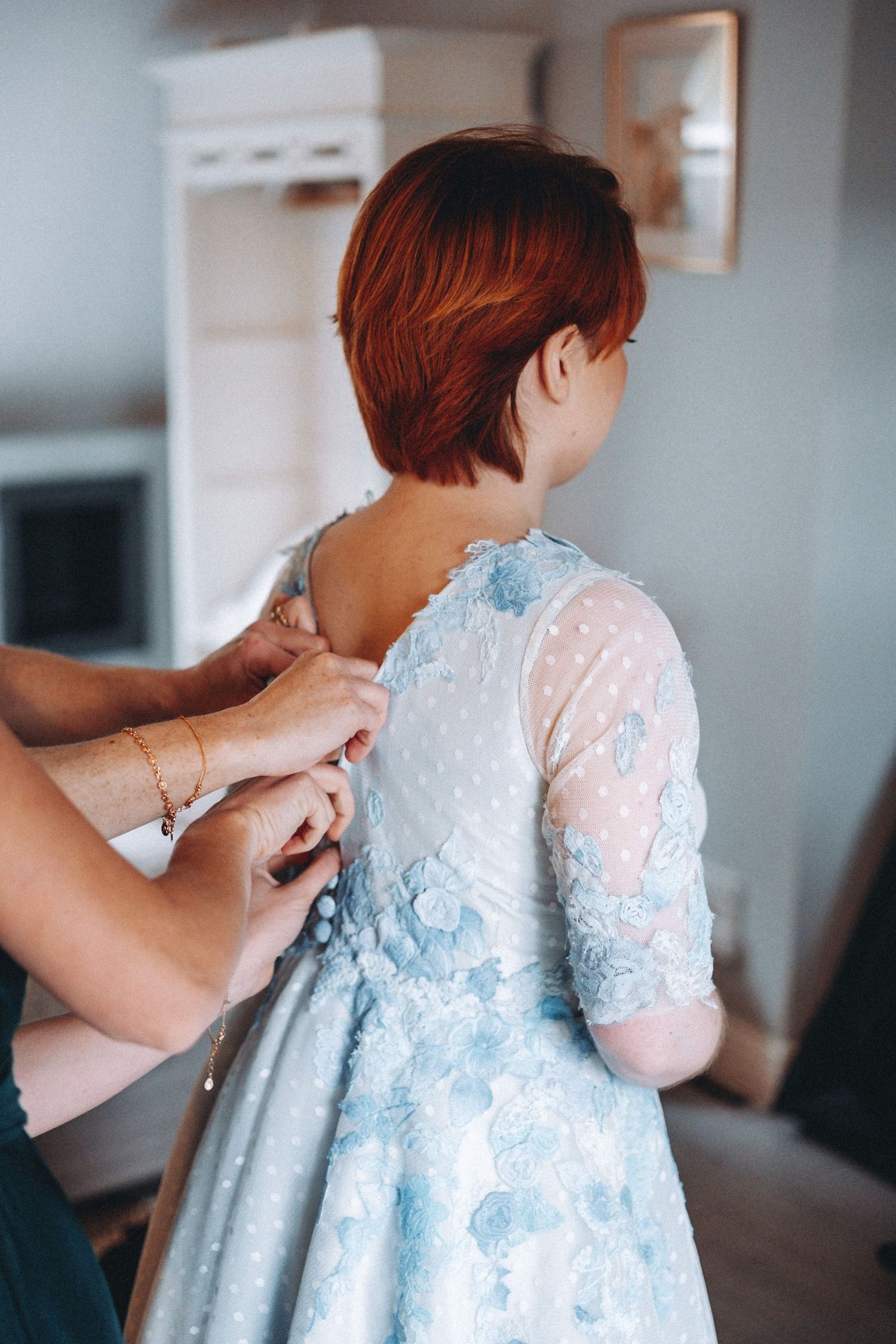 A close-up of hands fastening buttons on a light blue lace wedding dress.