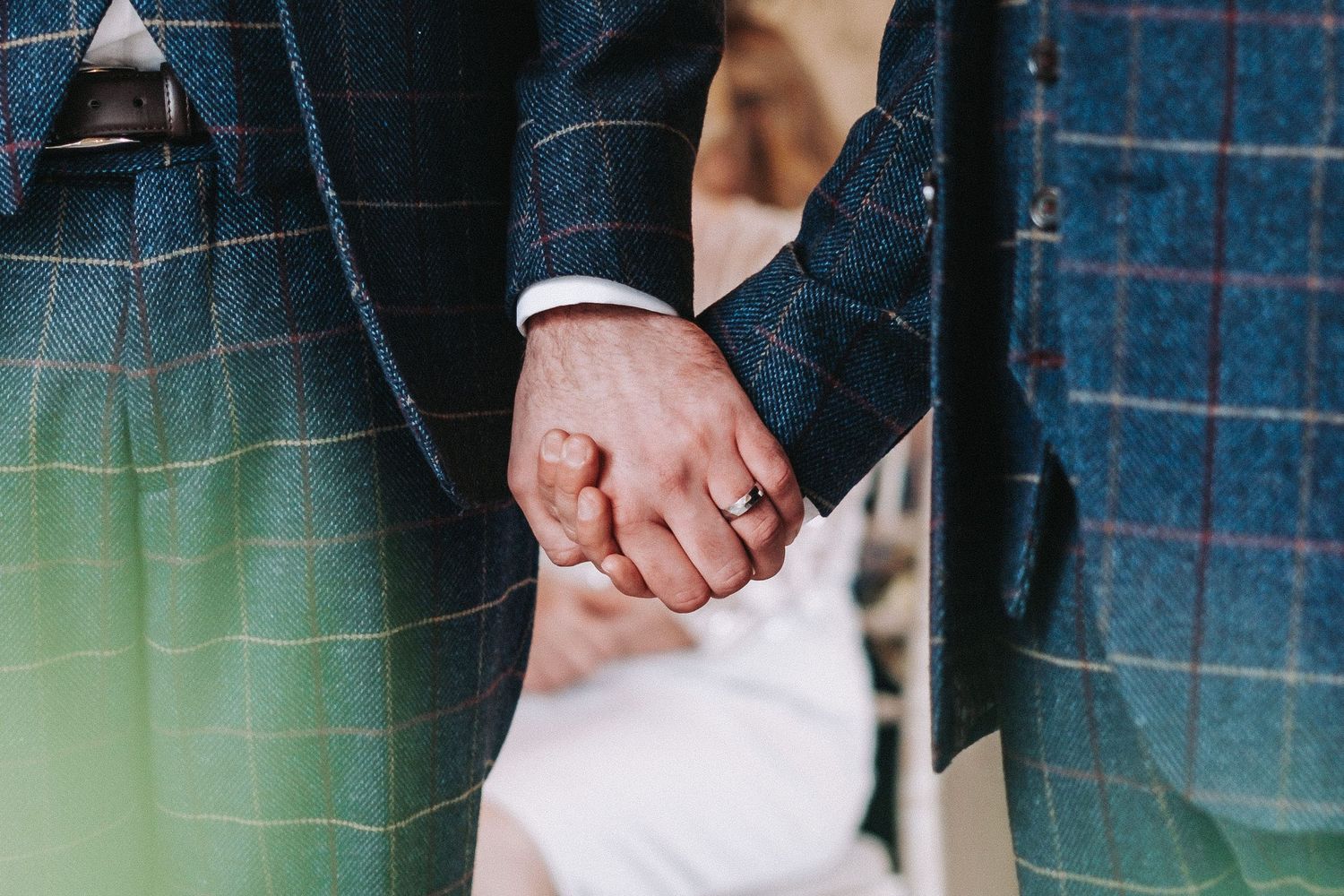 Wedding couple holds hands intimately while wearing elegant attire at an outdoor venue.