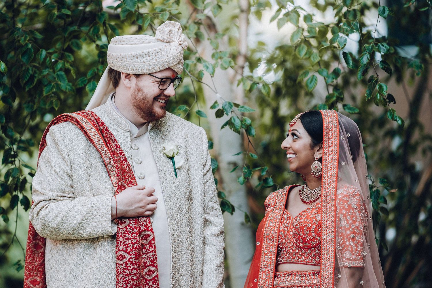 Guests in traditional Indian wedding attire gather outdoors among green foliage.