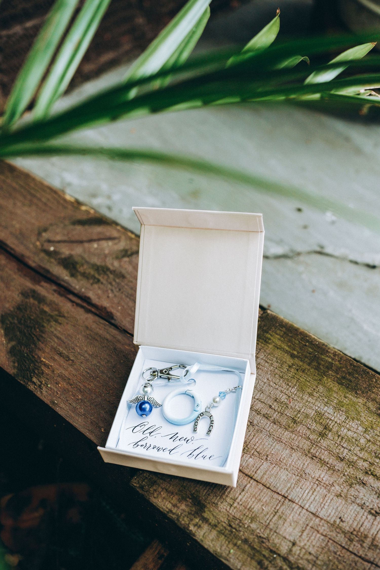 Wedding jewelry displayed in an open white box with blue and pearl accents on a rustic wooden surface near tropical leaves.