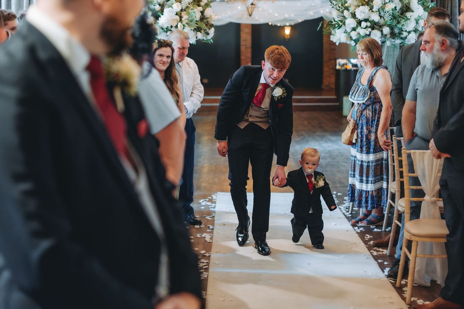 Two people walk down a church aisle at a formal event while guests look on from their seats.
