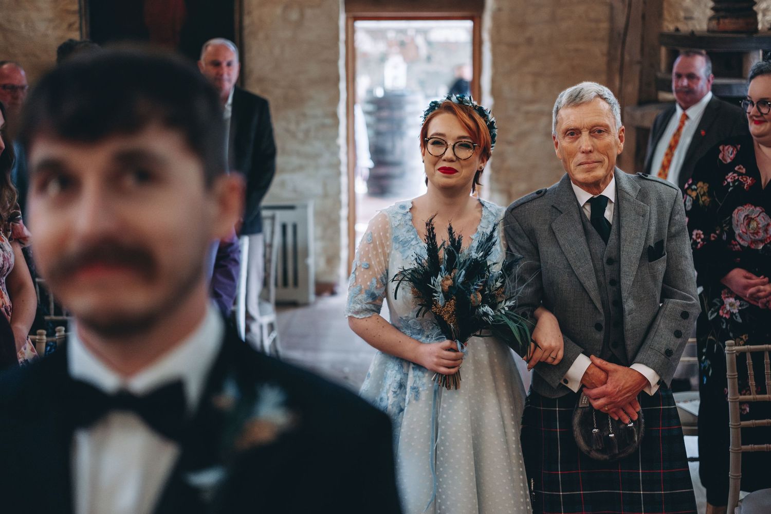 Bride and father walk down the aisle with the groom in black bow tie and suit, smiling in foreground.