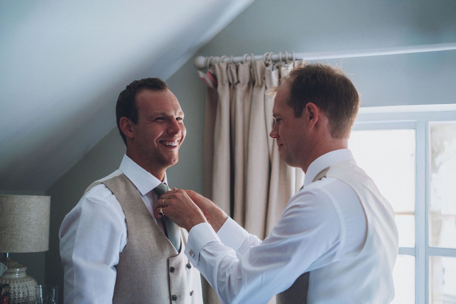 Two people helping each other with formal attire preparations in a sunlit room.