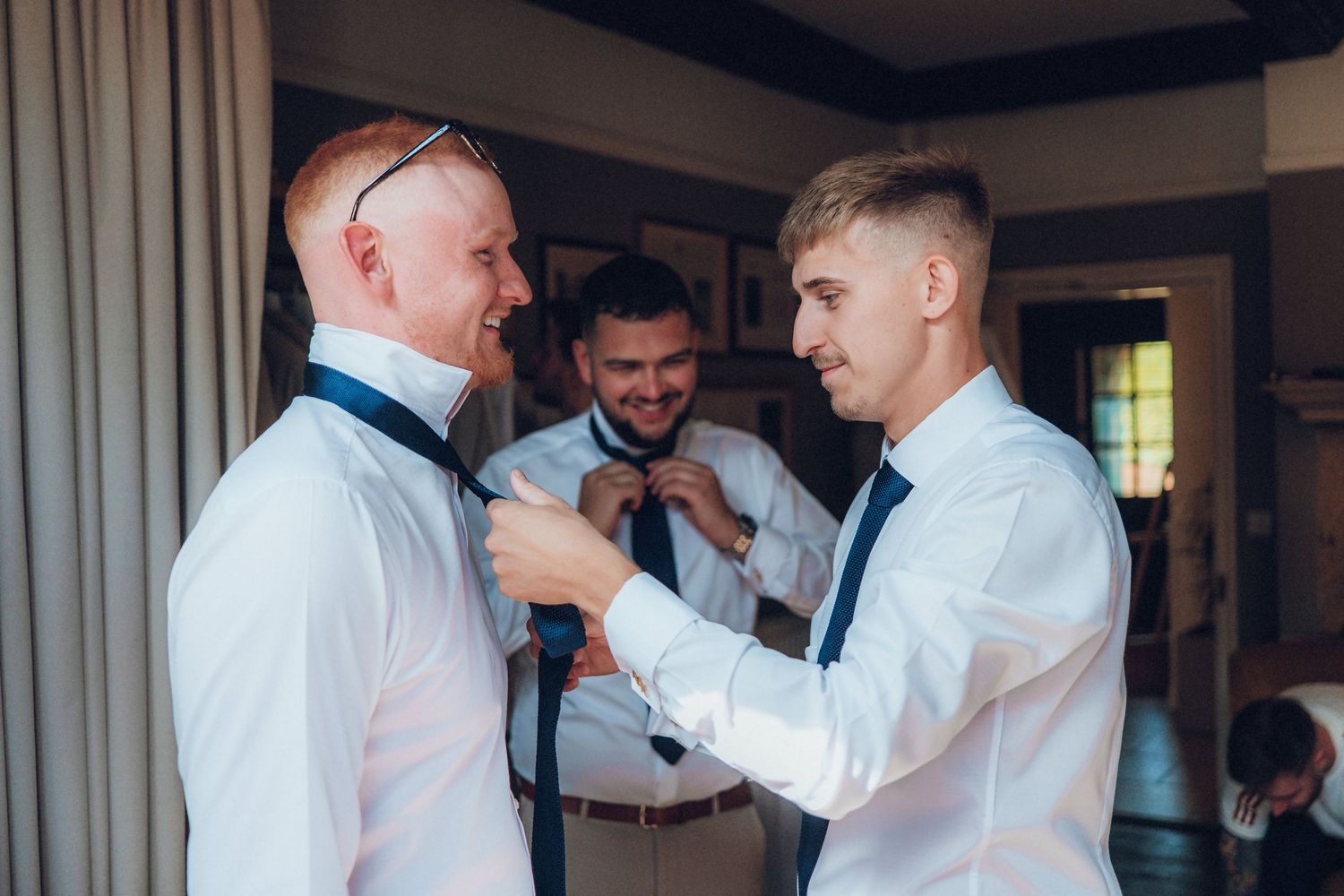 Groomsmen in white dress shirts and ties share emotional moments while getting ready for a wedding ceremony.