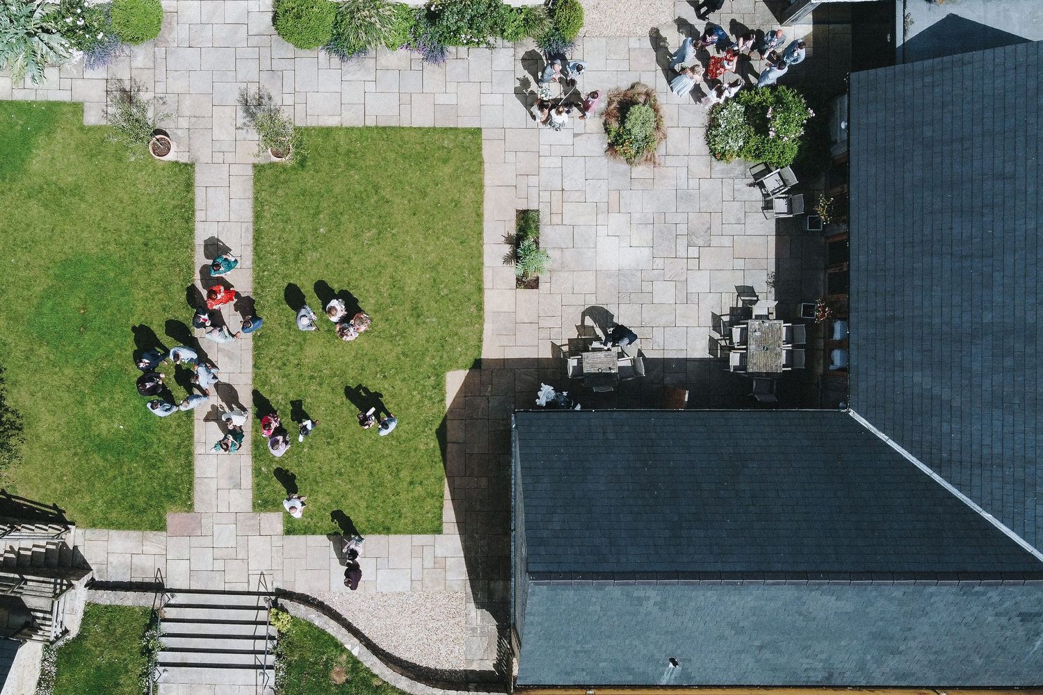 Aerial view of people gathering on stone paths and grass near a building with dark roofing.