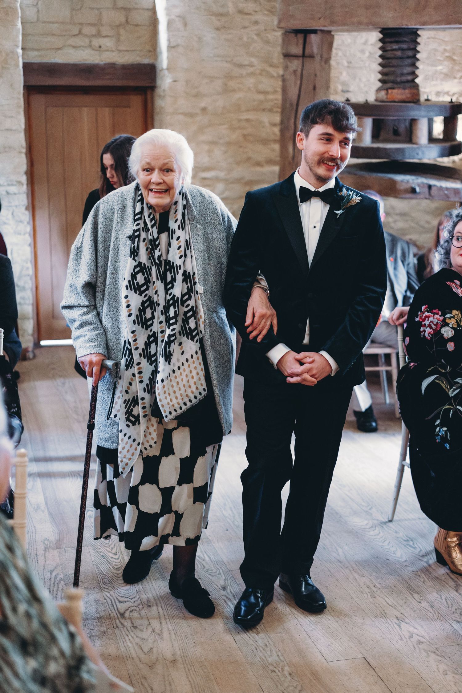 An elderly woman in a patterned dress stands next to a man in a tuxedo at a formal event.