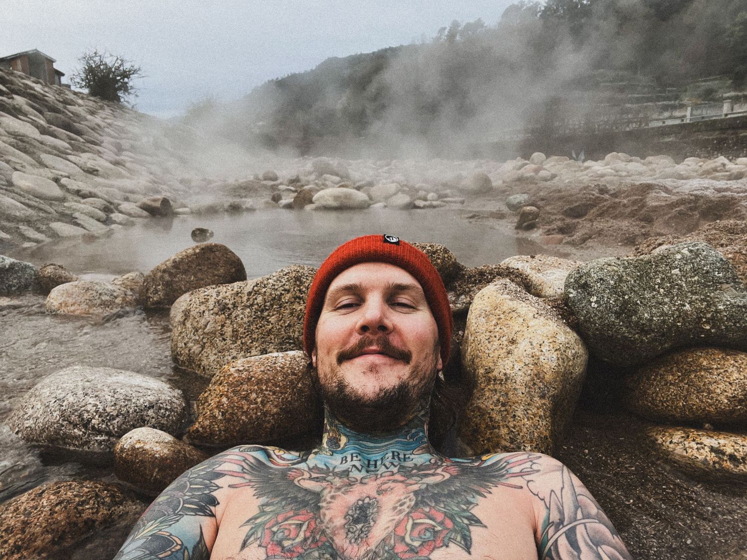 Person relaxing in steamy hot springs surrounded by large boulders wearing red beanie.