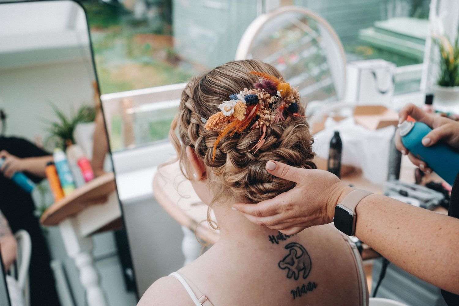 A stylist braids and arranges the brides intricate hairstyle with autumnal floral accessories, showing a back tattoo.