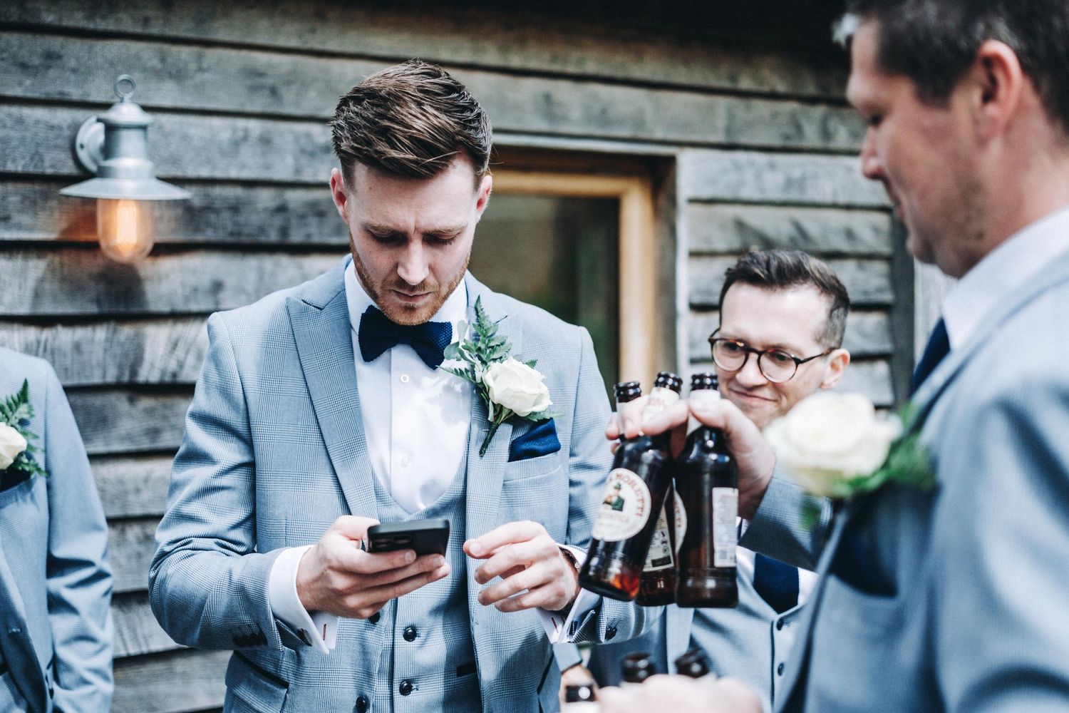 A wedding party in light gray suits gathers outdoors as people check their phones between formal photos.