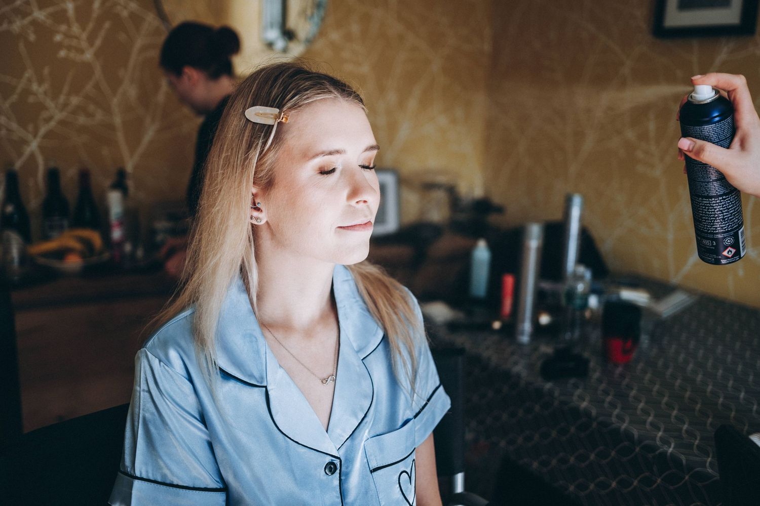 A person in a light blue blazer getting ready while someone holds a hair spray can in the background.