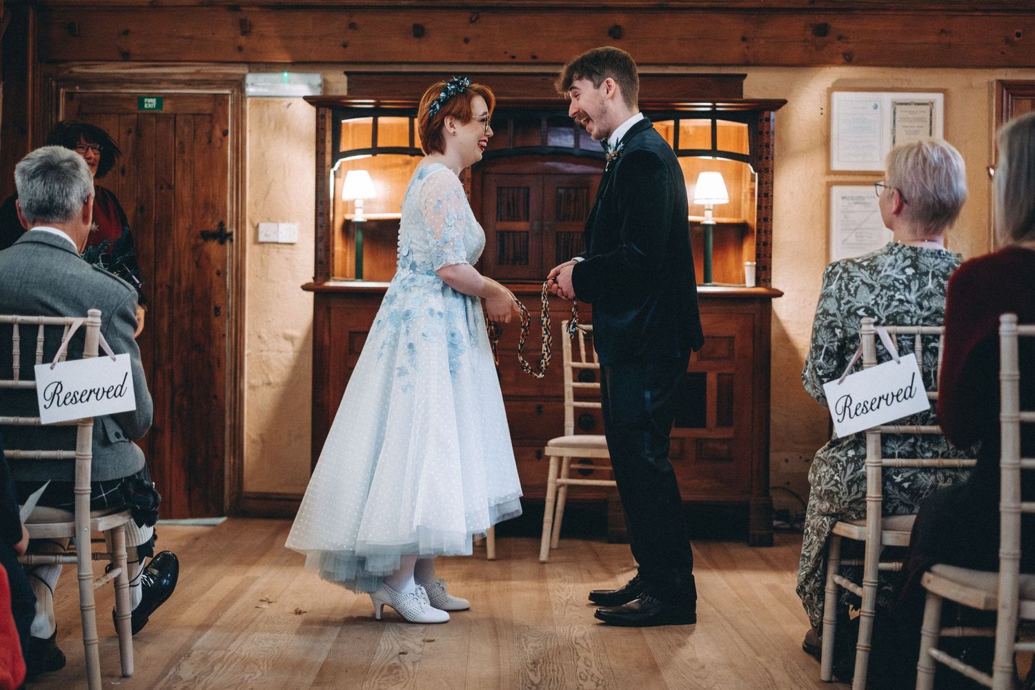 A couple tying the knot during a rustic indoor wedding ceremony with wooden floors and guests seated in chairs.