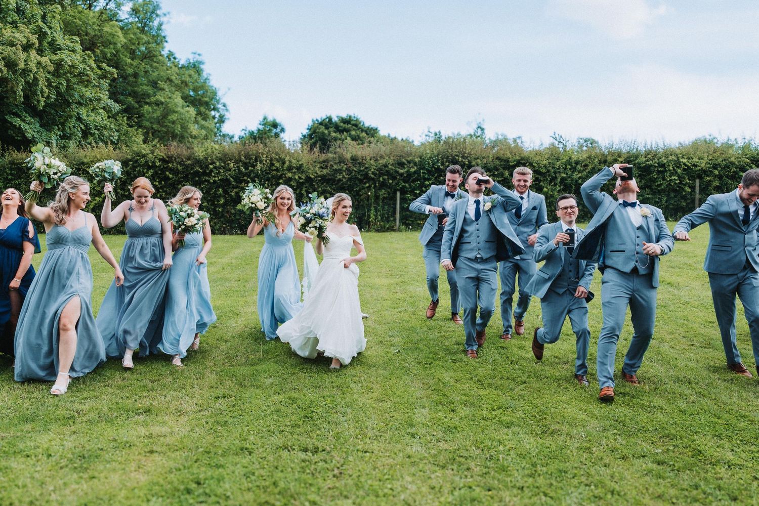 Wedding party in light blue attire runs joyfully across a green lawn during outdoor celebration.