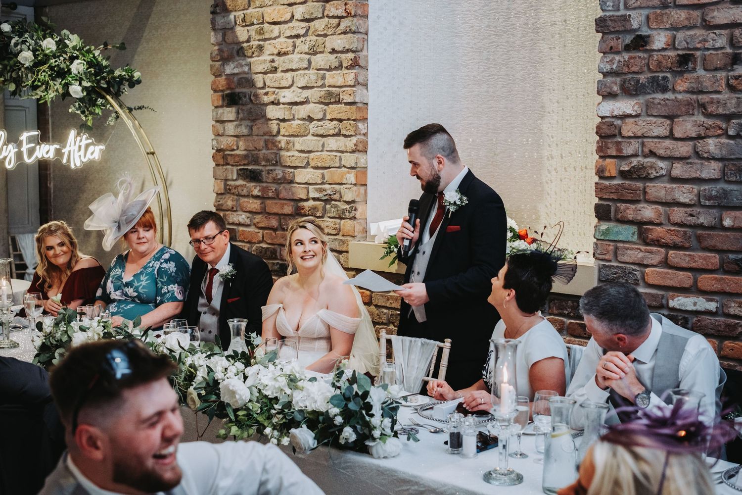 Guests seated at an elegant wedding reception table with brick walls and floral decorations.