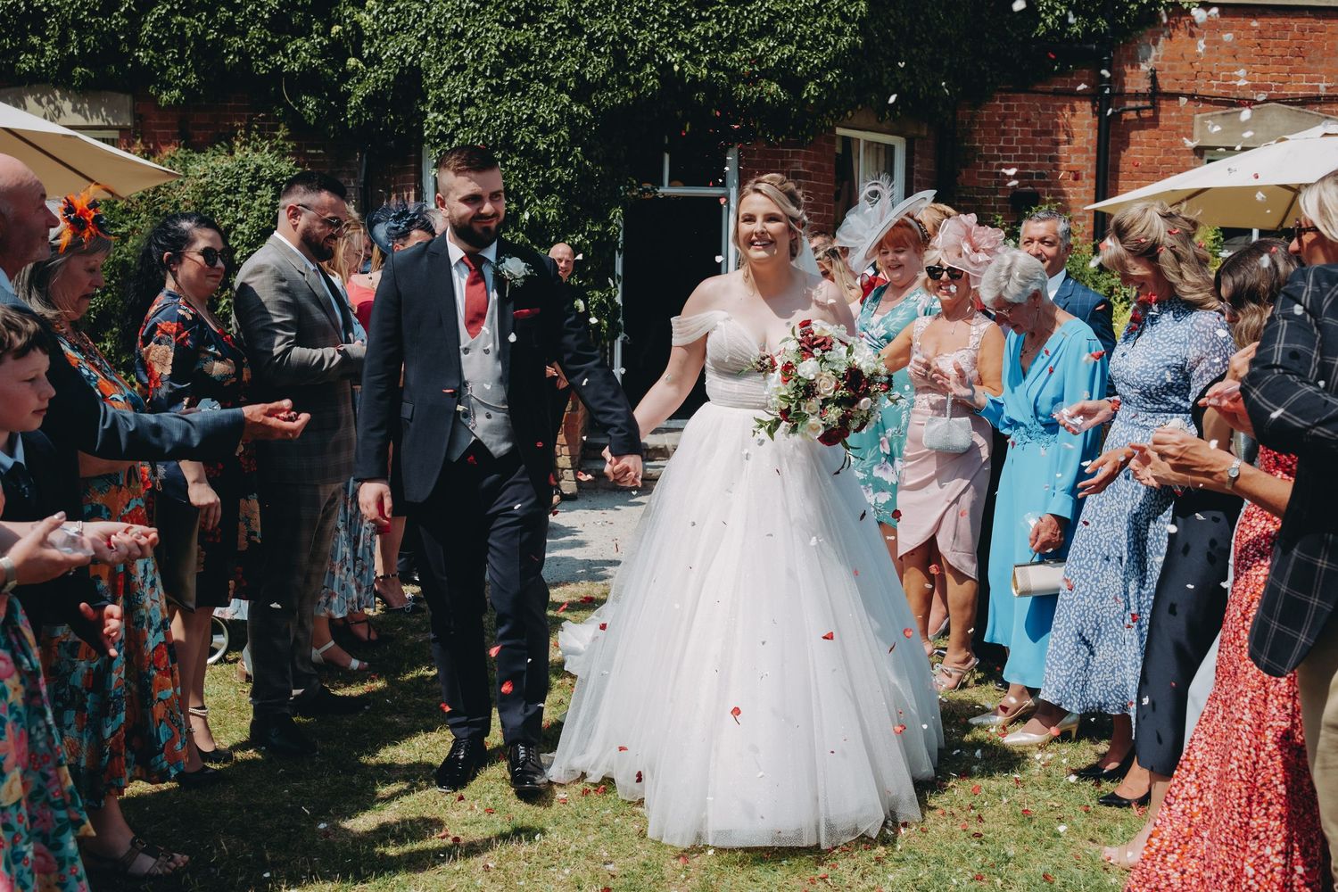 Wedding guests throwing confetti over newlyweds on a sunny garden lawn during outdoor celebration.