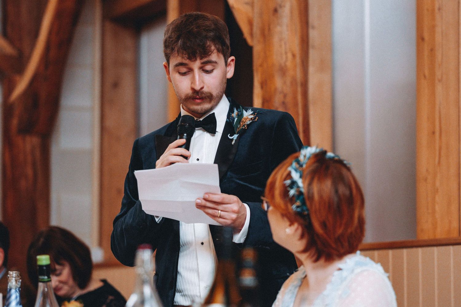 A groom in a black tuxedo delivers a wedding speech while holding papers at the reception.