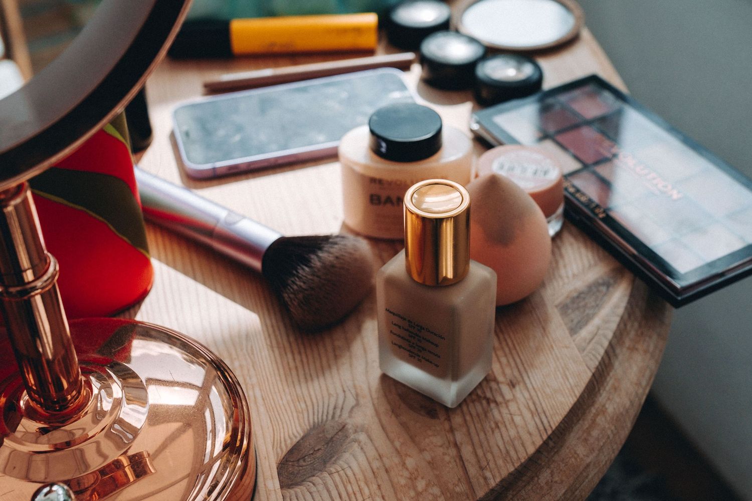 Makeup products including foundation bottle, brushes, and cosmetic palettes arranged on wooden surface with natural lighting.