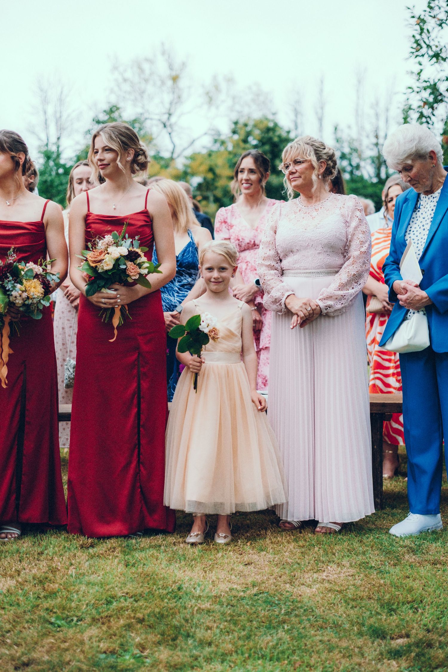 Wedding party in red and pink dresses posing together outdoors with flower bouquets.