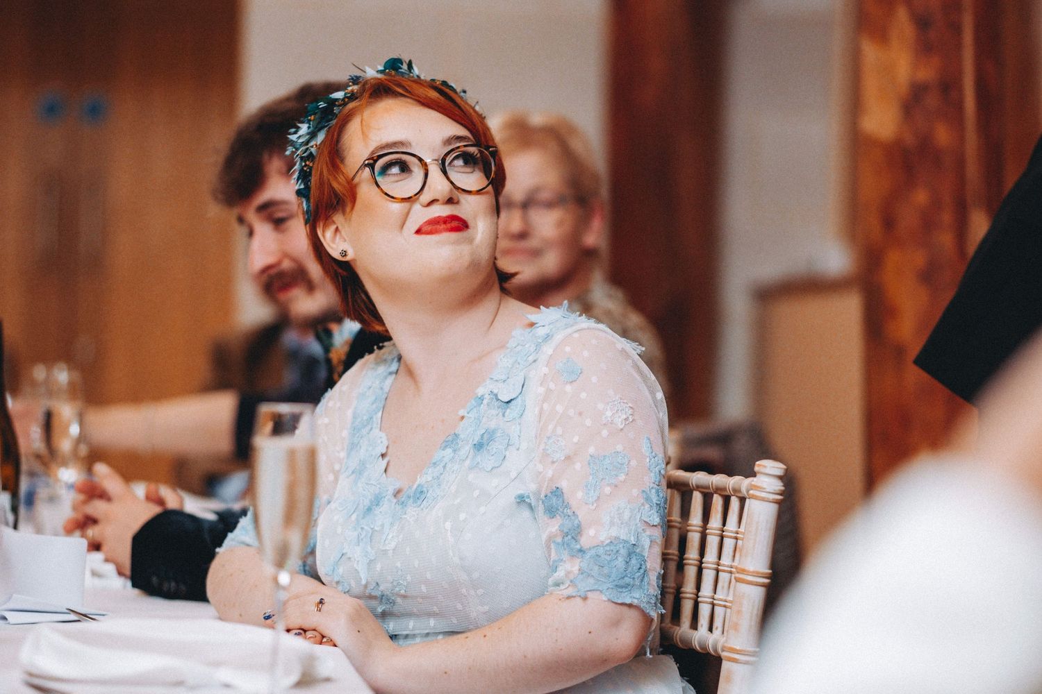 Reception guest in a light blue dress and vintage glasses listening intently during celebrations.
