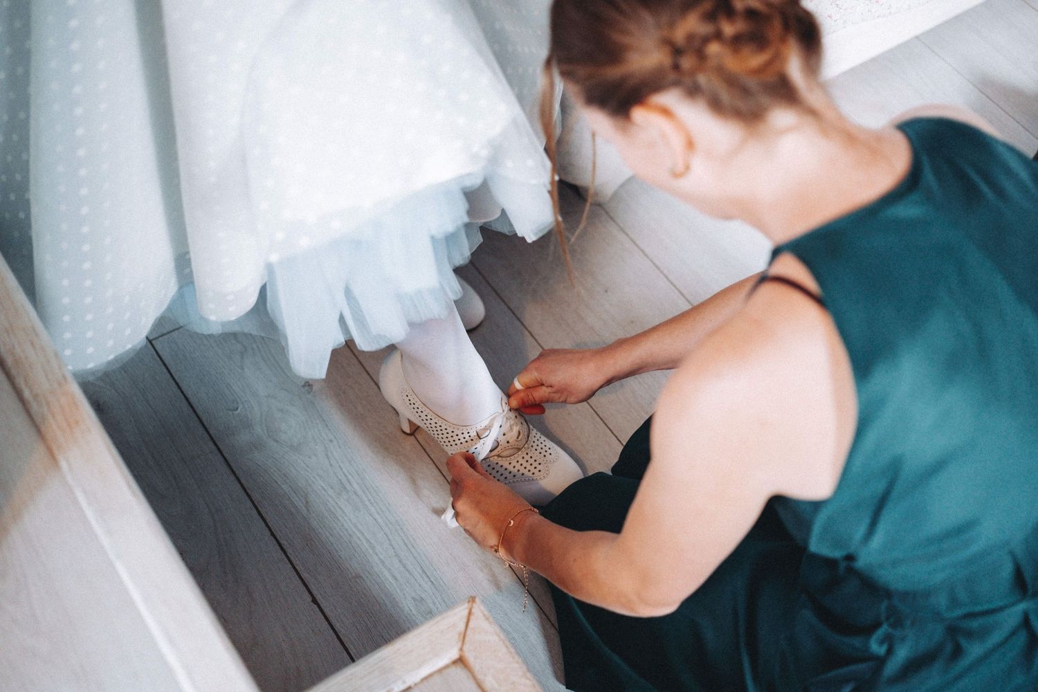 Bridesmaid in an emerald green dress helps Bride put on delicate footwear in the Ryder Barn at Owlpen Manor..