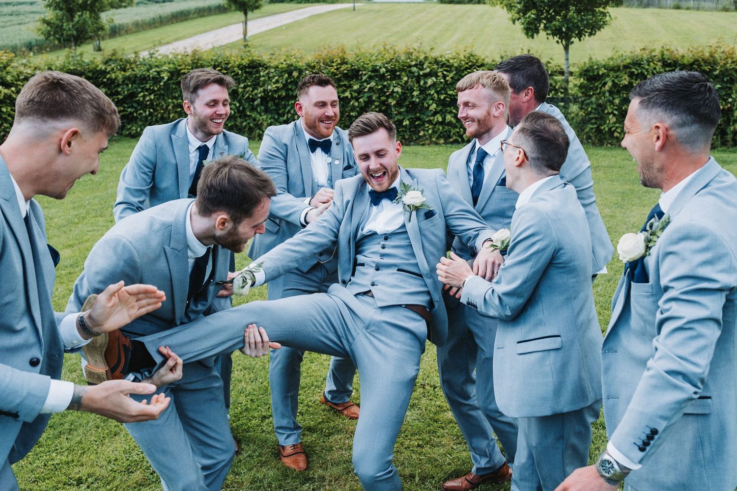 Groomsmen in light blue suits laughing and posing together on a garden lawn.