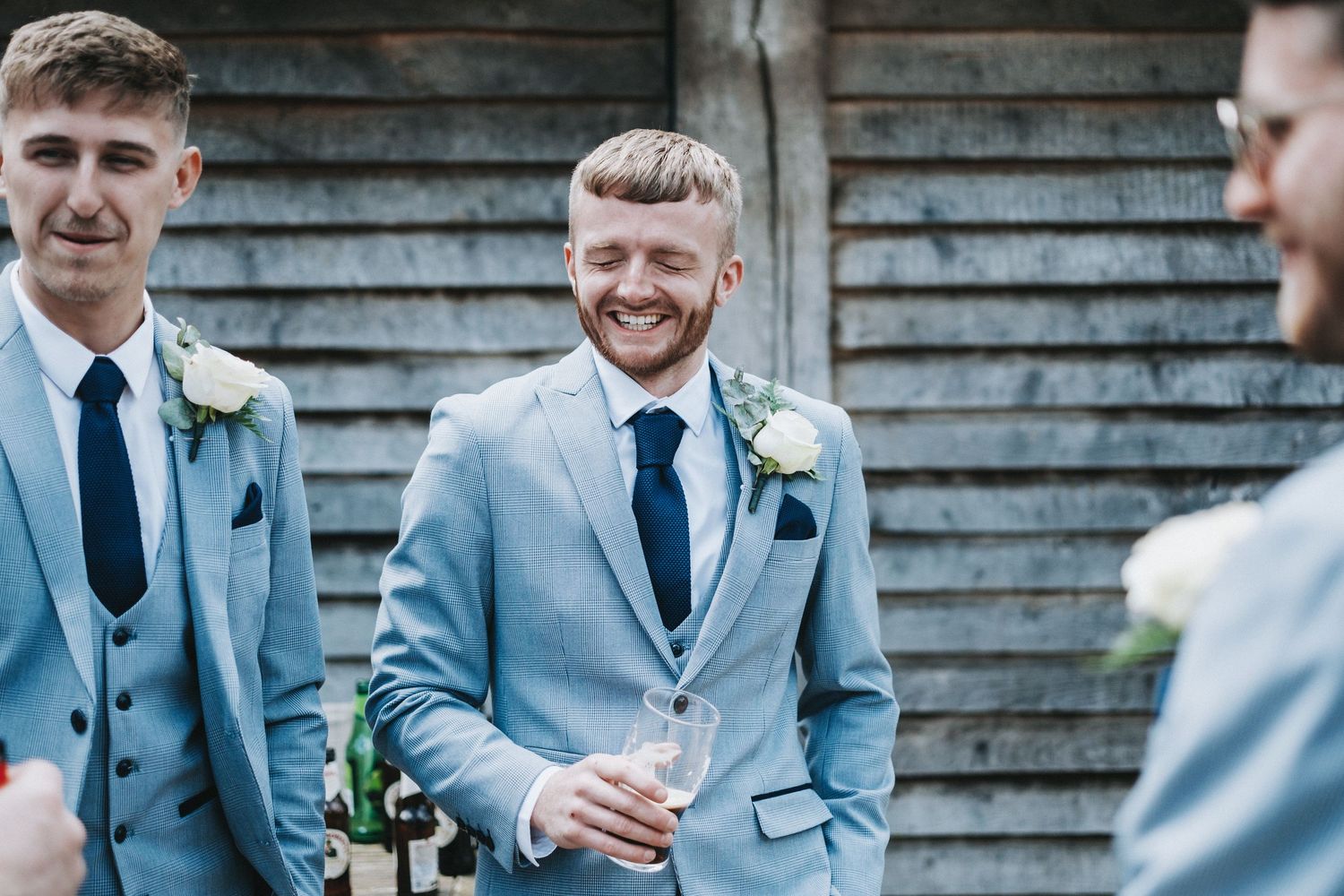 Wedding party members in light blue suits and navy ties share laughs during outdoor celebration.