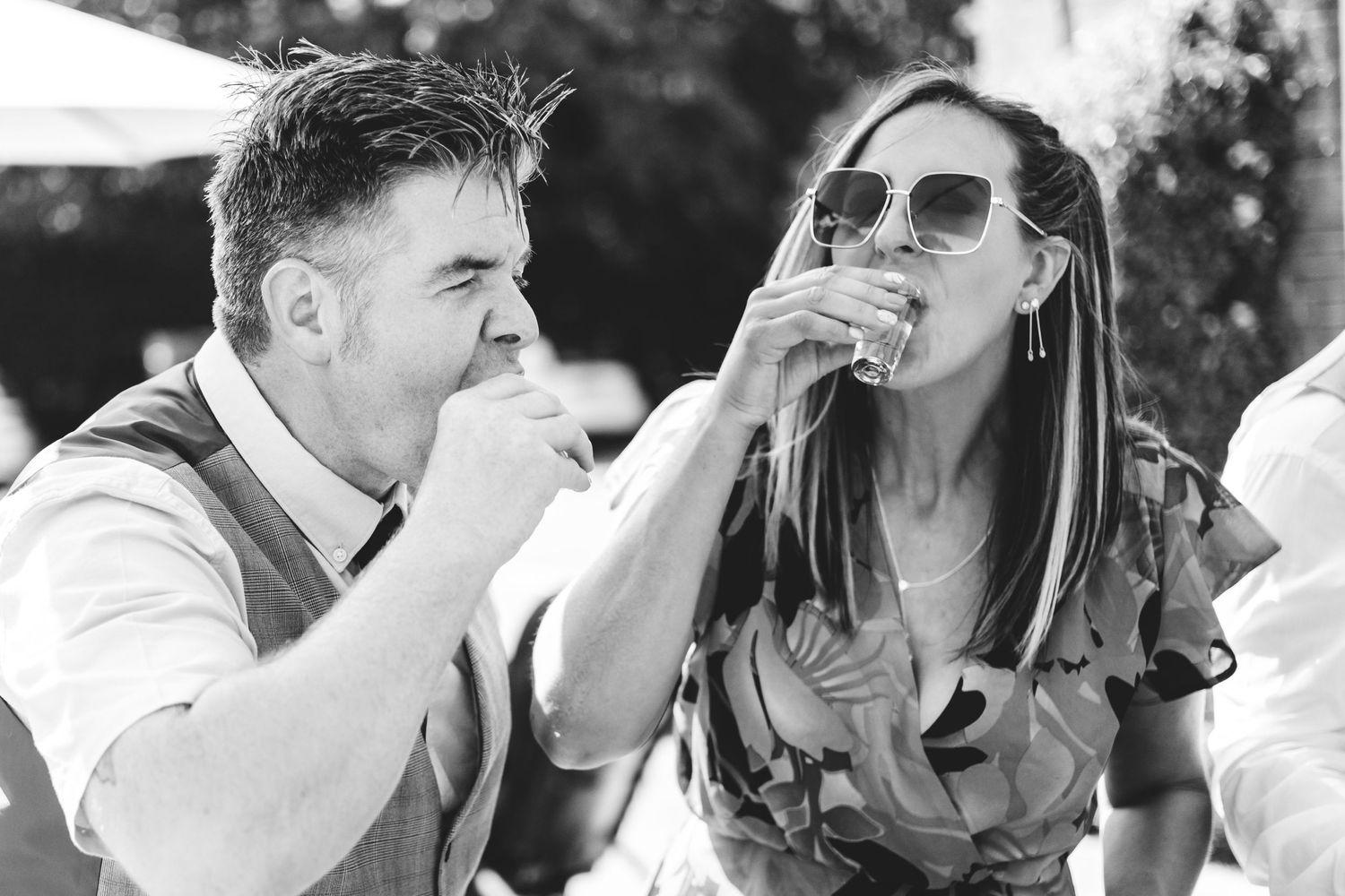 A black and white photo of two people sharing drinks and laughing together at a wedding.
