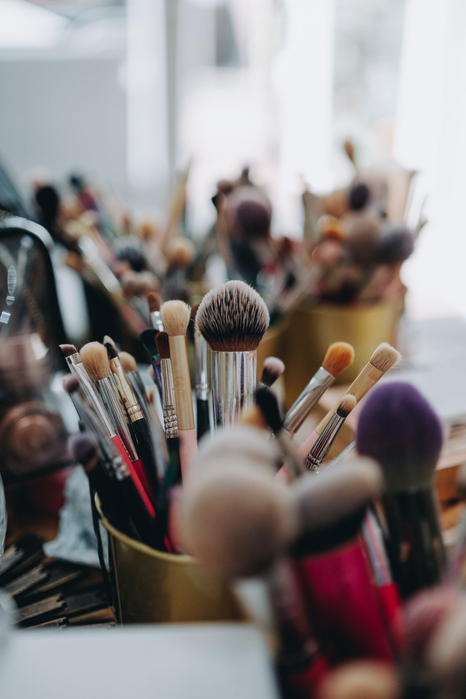 Collection of makeup and cosmetic brushes arranged in gold holders on white surface in soft focus.