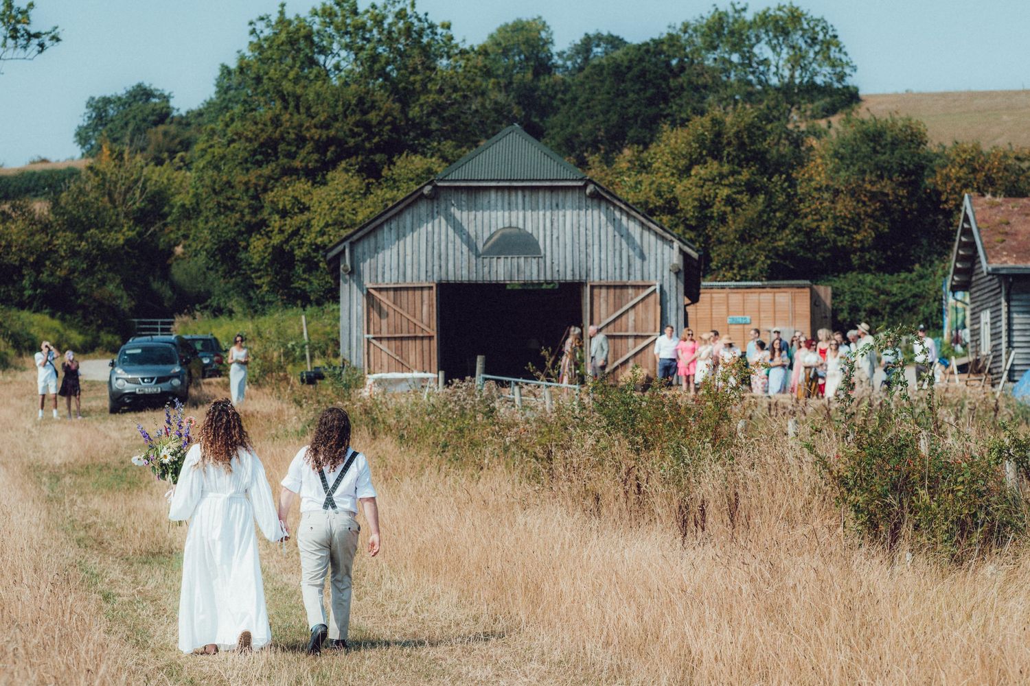 People in white clothing walking through a grassy field near a barn on a sunny day.