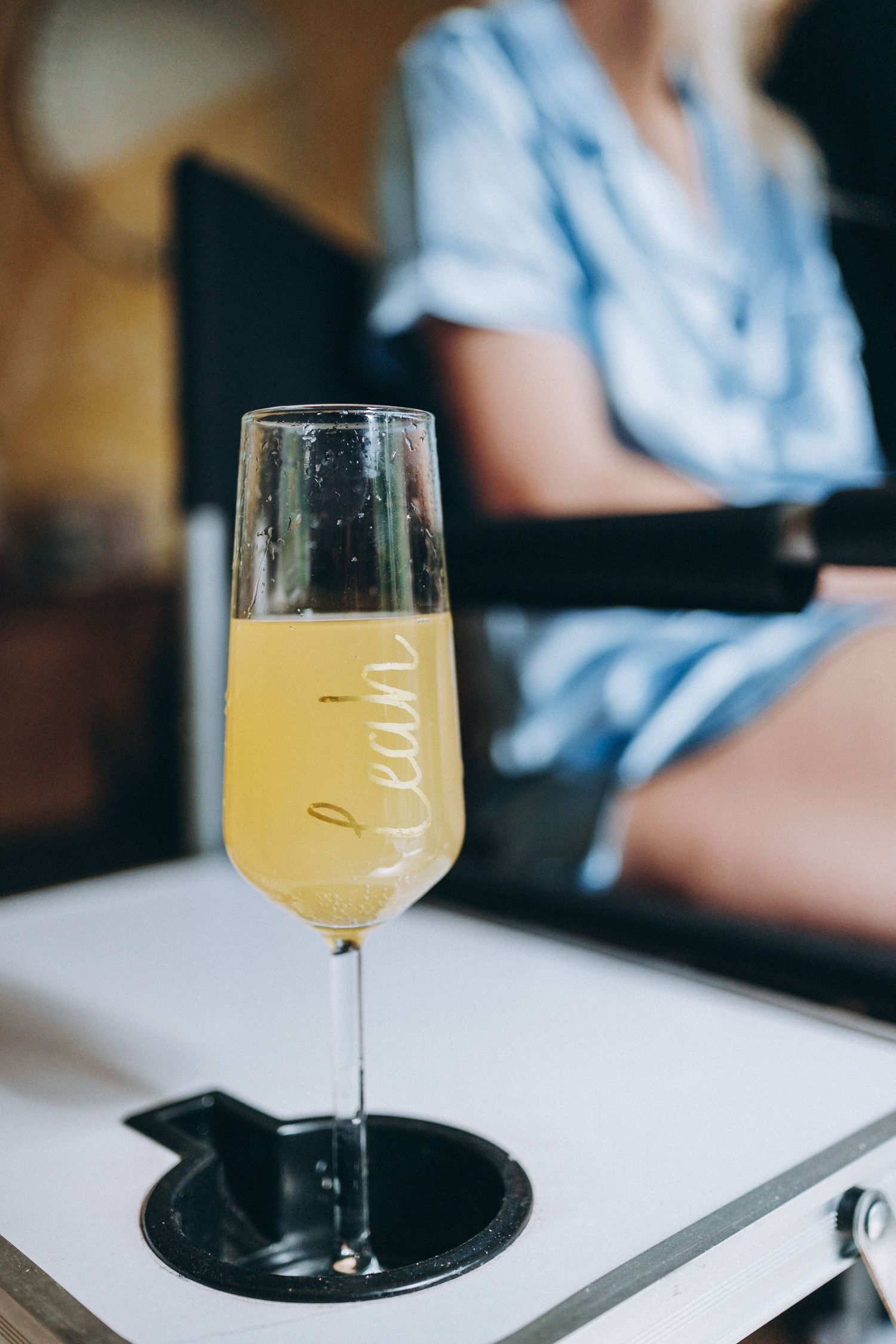 A glass of golden mimosa cocktail sits on a white surface with someone wearing blue in the background.