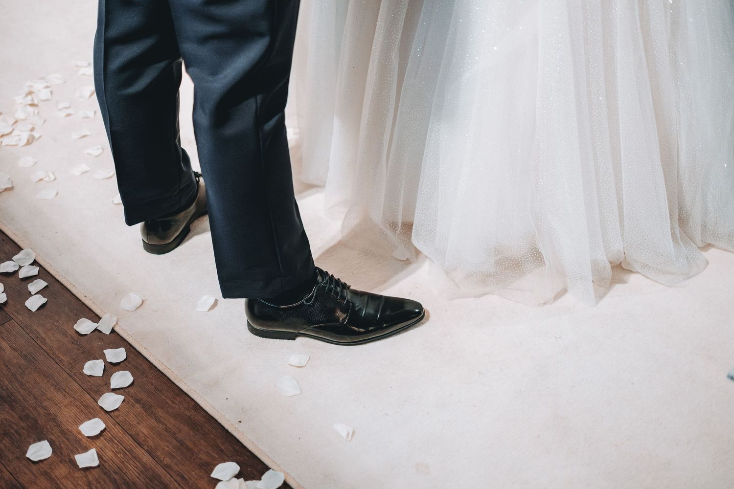 Black dress shoes next to a flowing white wedding dress hem with scattered rose petals on dark wooden floor.