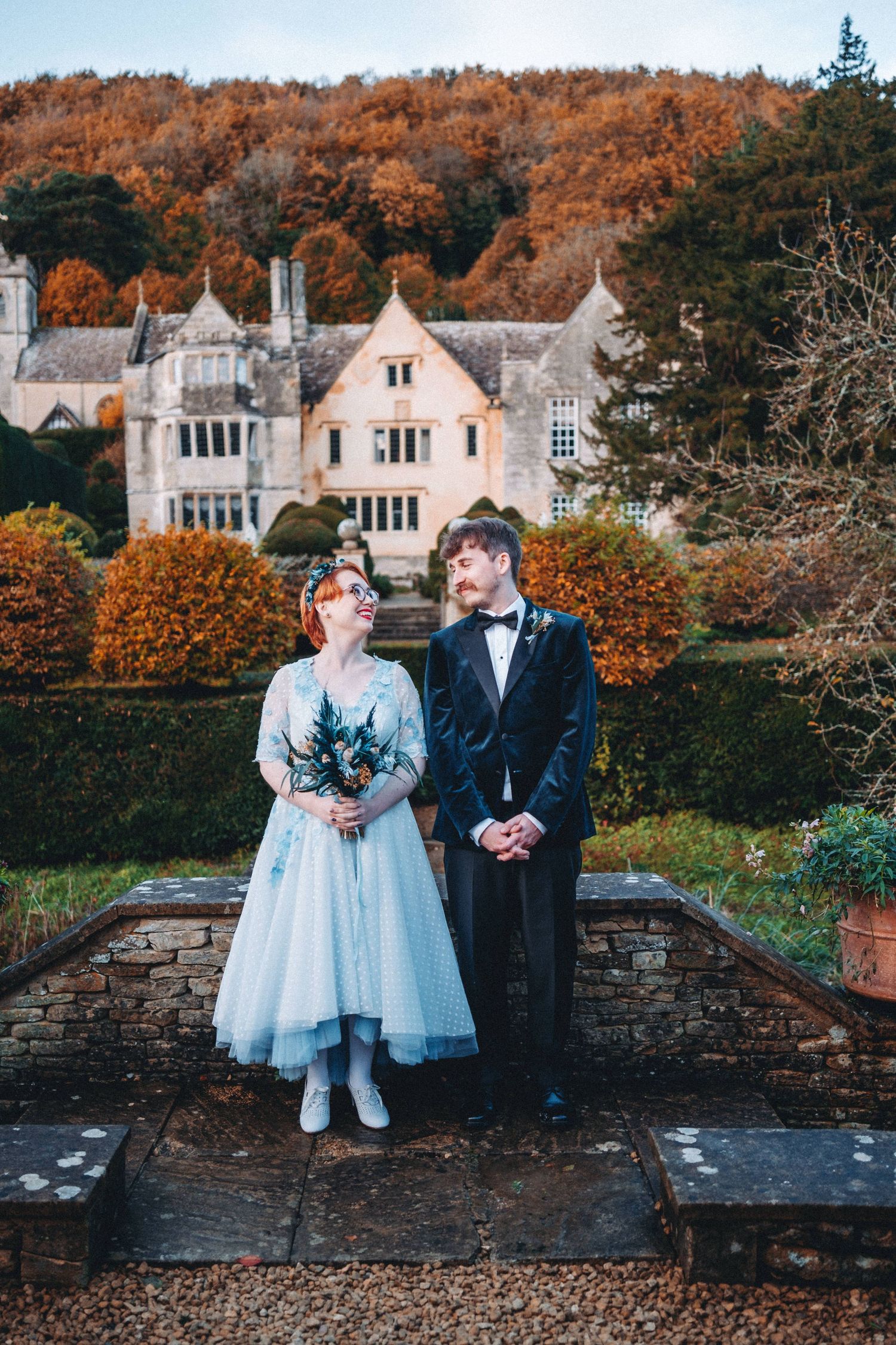 Newlyweds pose in front of grand stone mansion with autumn foliage backdrop.