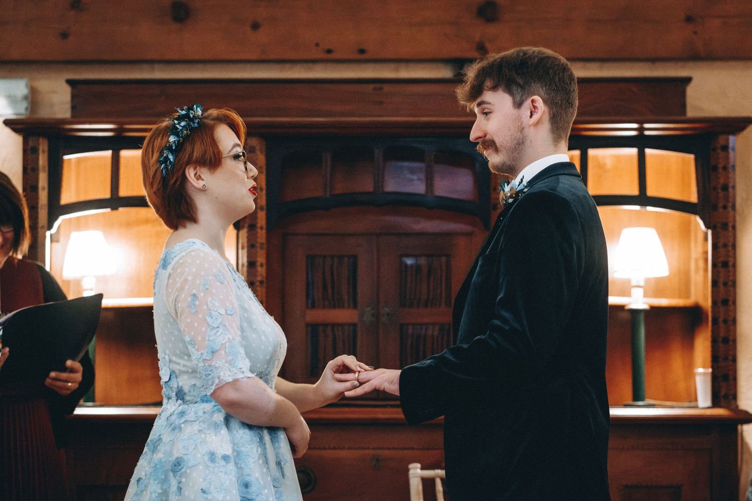A couple in wedding attire exchange rings during a wedding ceremony in a warmly lit room with wooden decor.