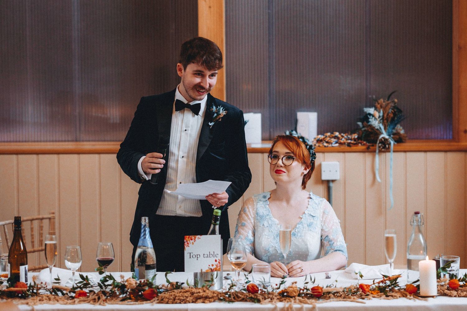 A couple at their wedding reception head table during a speech moment, decorated with floral centerpieces.
