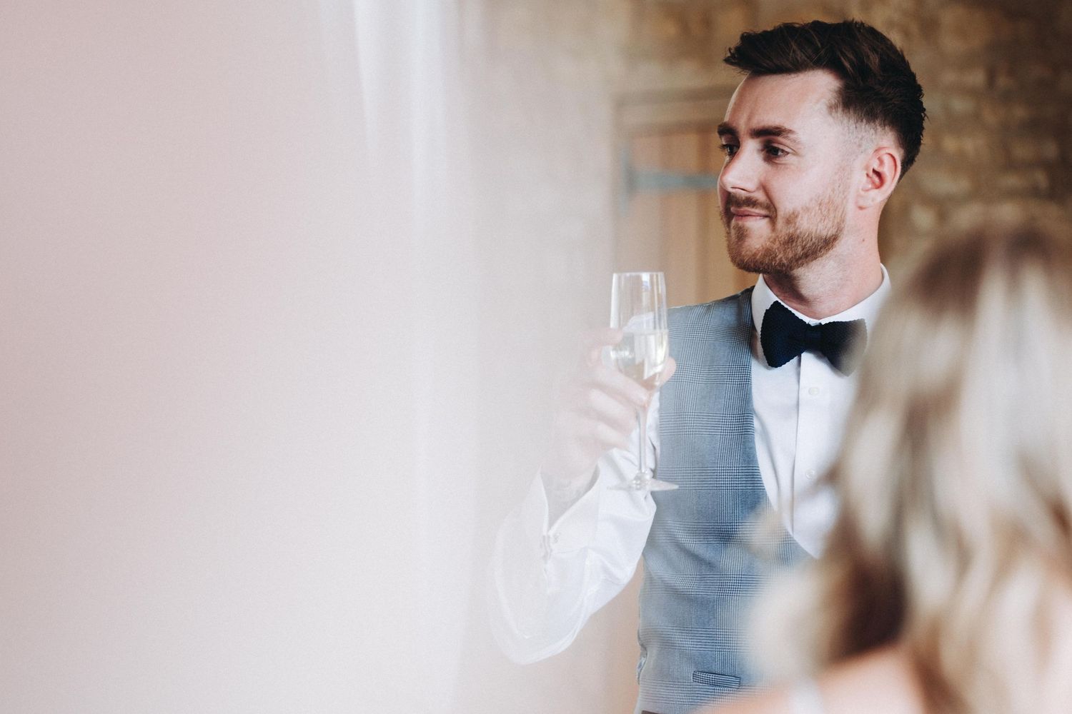 A well-dressed person in a gray vest and bowtie raises a champagne glass while celebrating at a formal event.