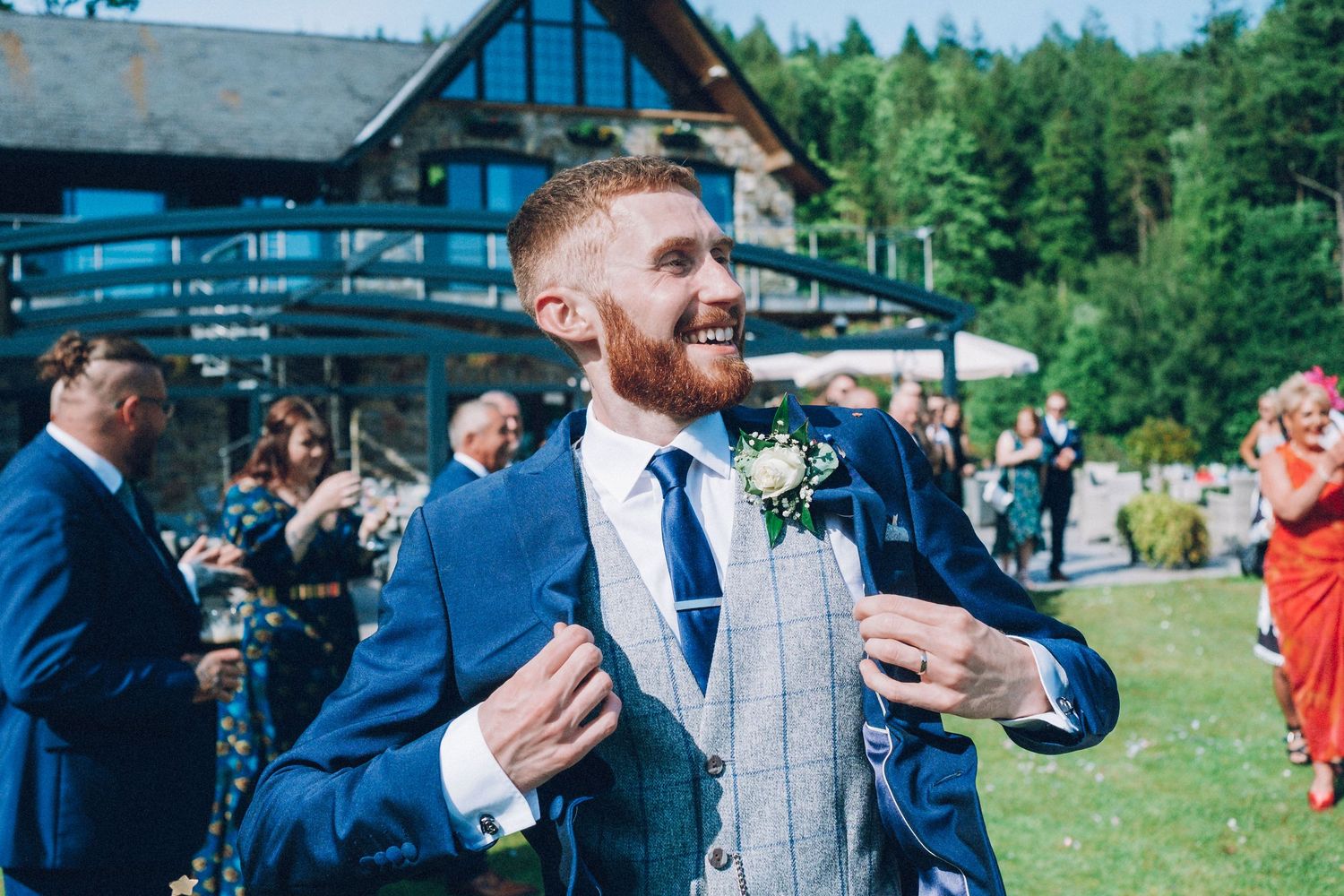 A wedding guest in a blue suit and gray vest smiles during an outdoor celebration.