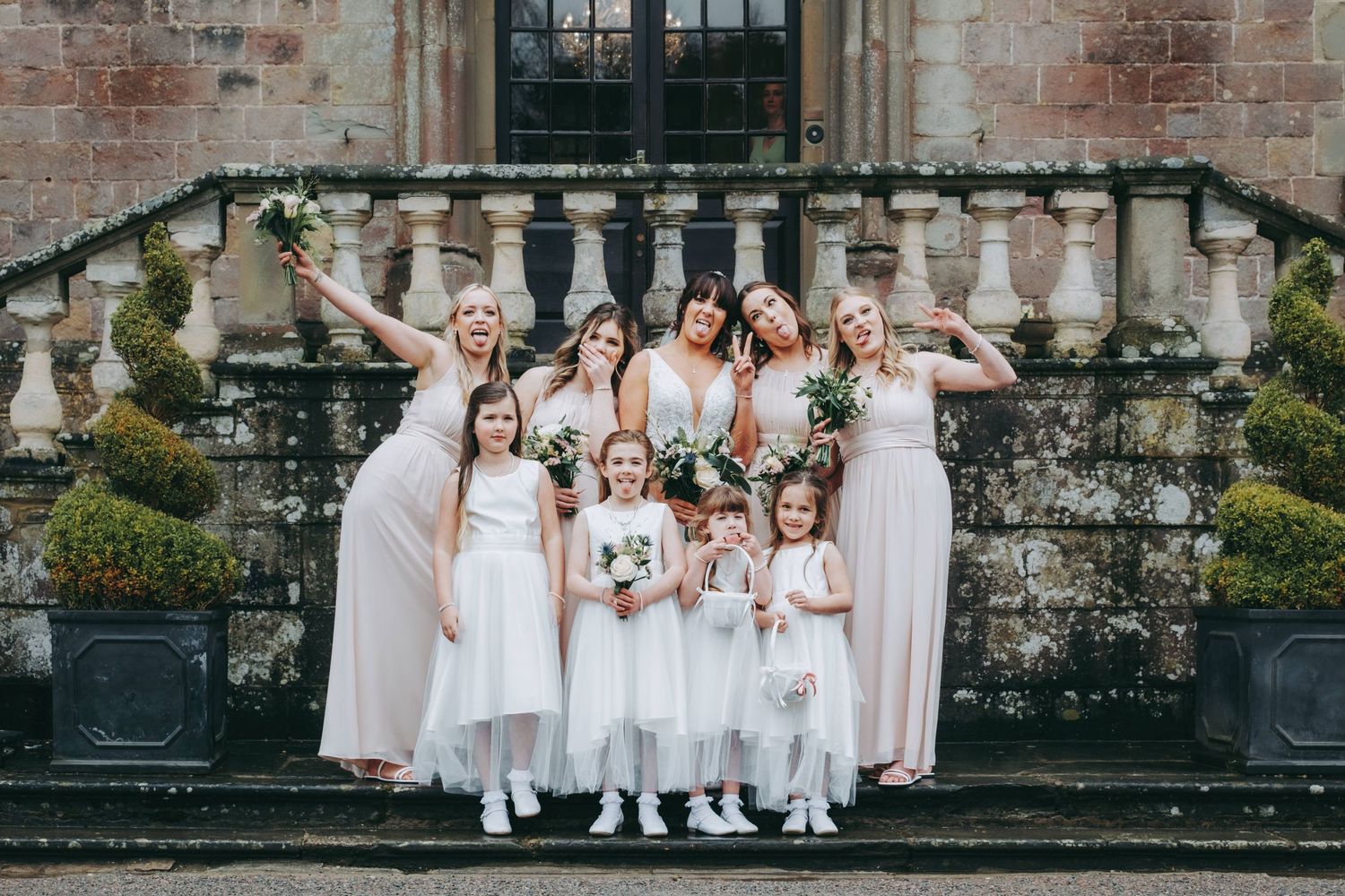 A wedding party of bridesmaids and flower girls in soft pink dresses pose on stone steps against a historic brick wall.