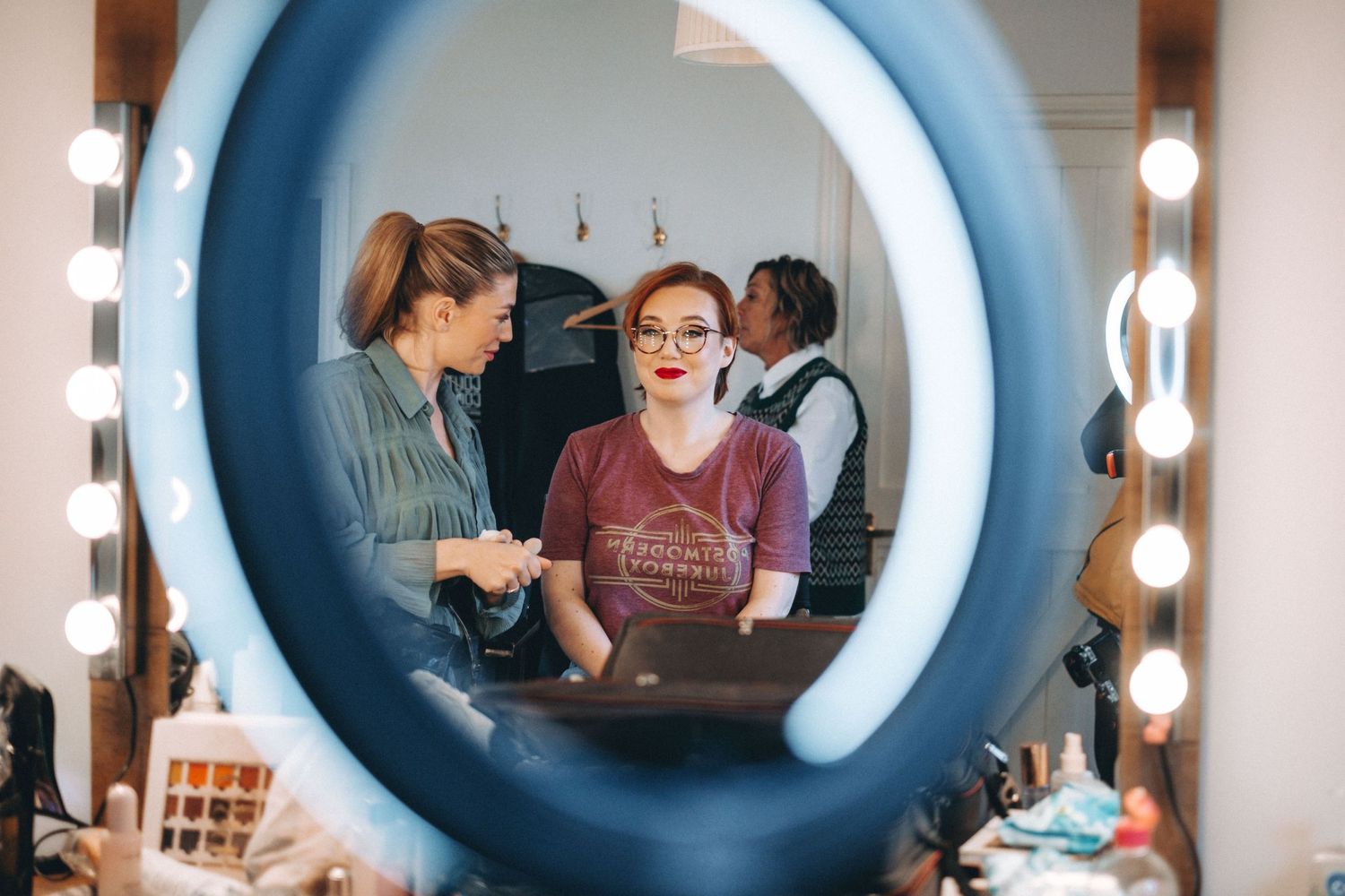 Behind-the-scenes view through a lighted makeup mirror showing preparation for an event.