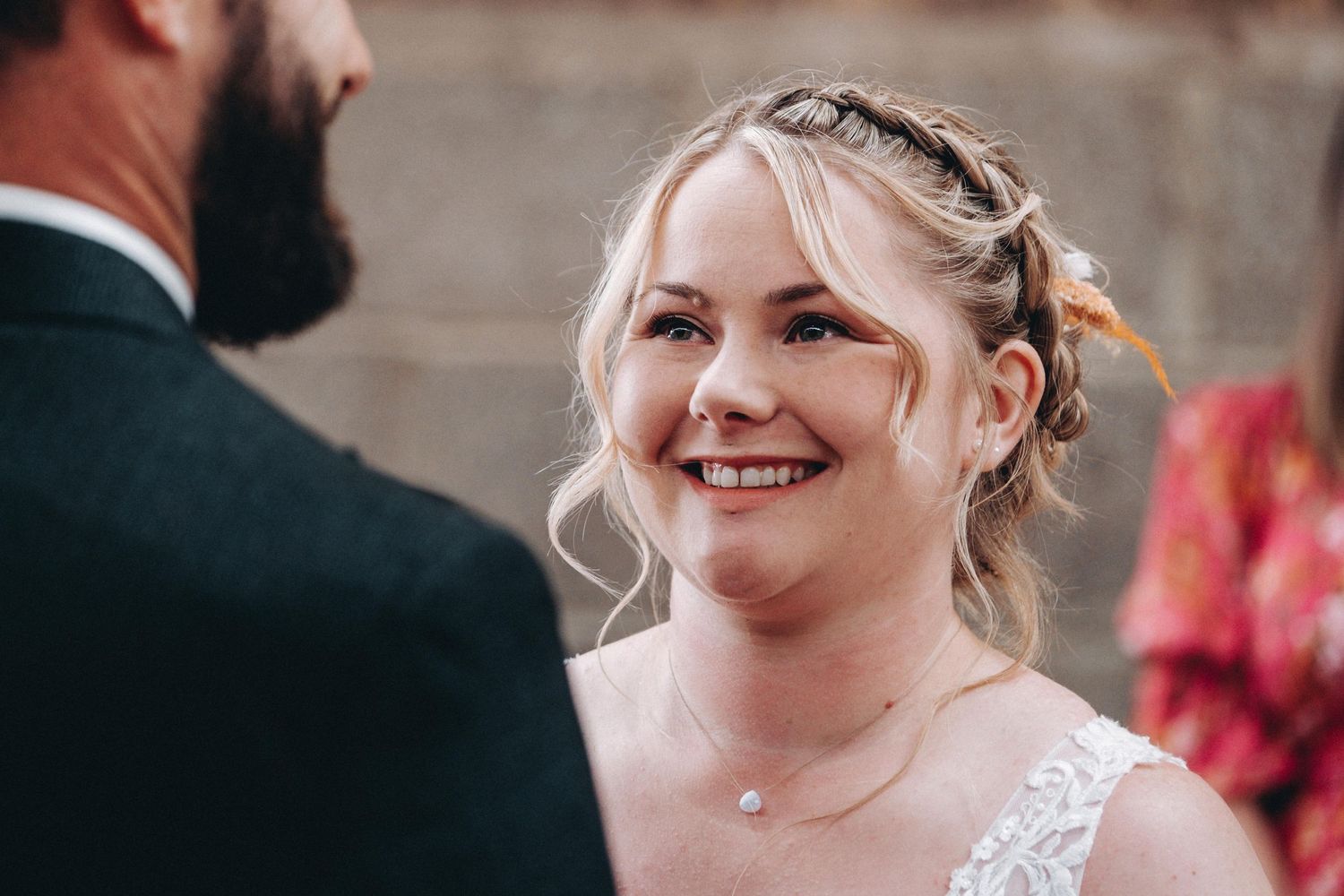 A bride with braided hair and a lace wedding dress smiles joyfully during her wedding ceremony.