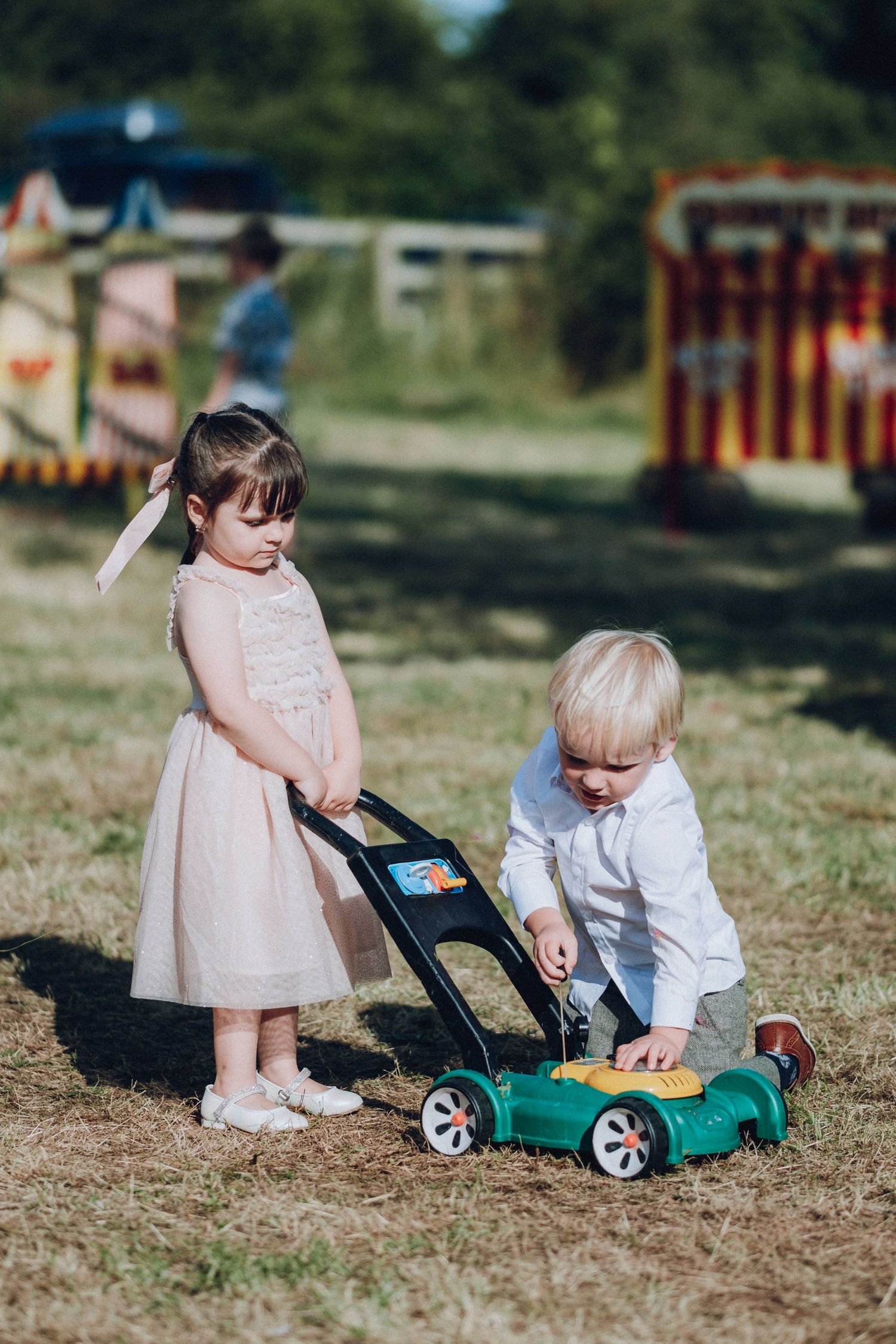 Children playing with a toy lawn mower on grass during an outdoor celebration.