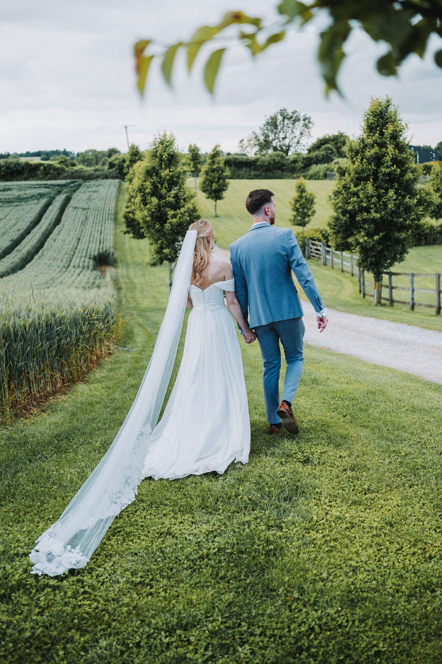 A couple in wedding attire walks through a scenic countryside field with tall grass and trees.