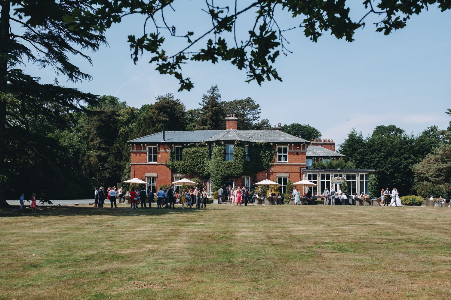 Large brick country house with manicured lawn and guests gathering for outdoor event.