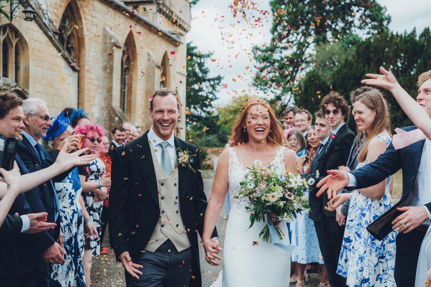 Newlyweds walking through shower of confetti outside historic church building.