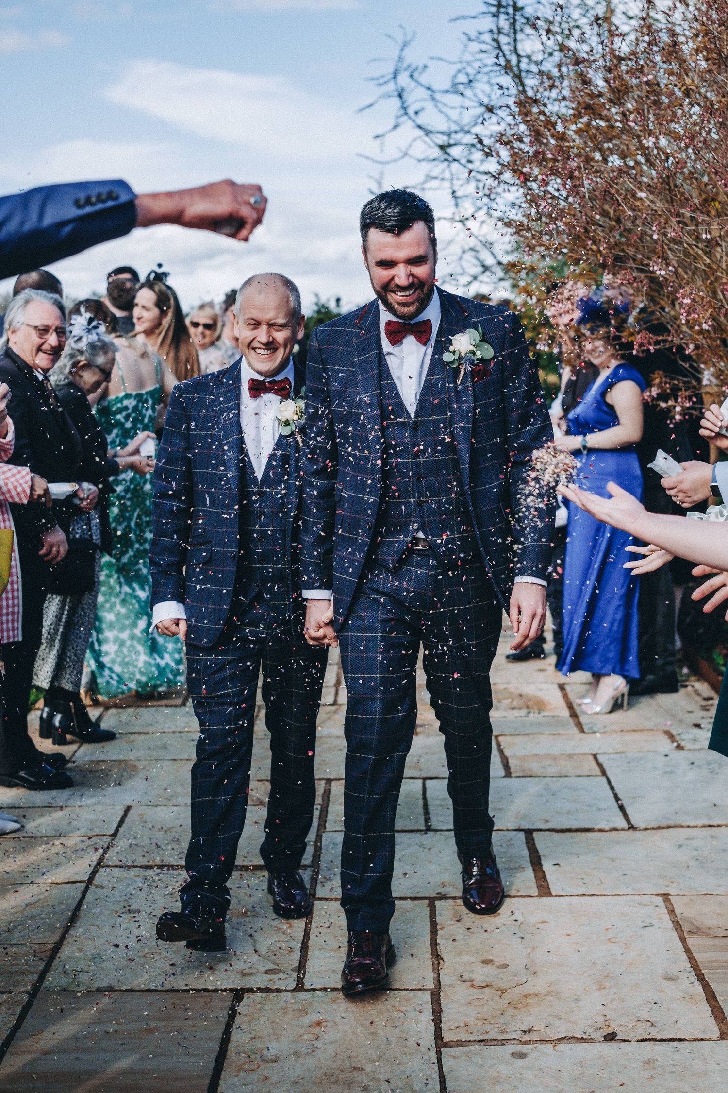 Wedding guests throw confetti over formally dressed people on stone pathway.